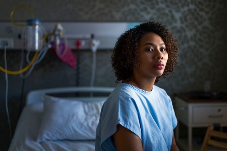 A woman looking pensively while wearing a hospital gown sitting on a hospital bed in a dimly lit room