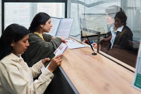 Two young adult Hispanic women standing at visa center counter holding documents and passports, engaging in conversation while waiting for service, female staff member visible in background